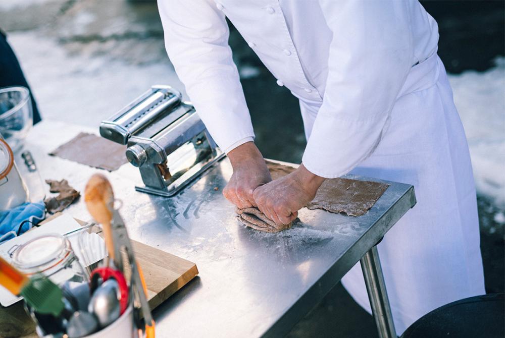 Un chef en uniforme blanc p&eacute;trit de la p&acirc;te sur une table en m&eacute;tal farin&eacute;e, avec une machine &agrave; p&acirc;tes &agrave; proximit&eacute;. Des ustensiles et un bocal sont visibles.