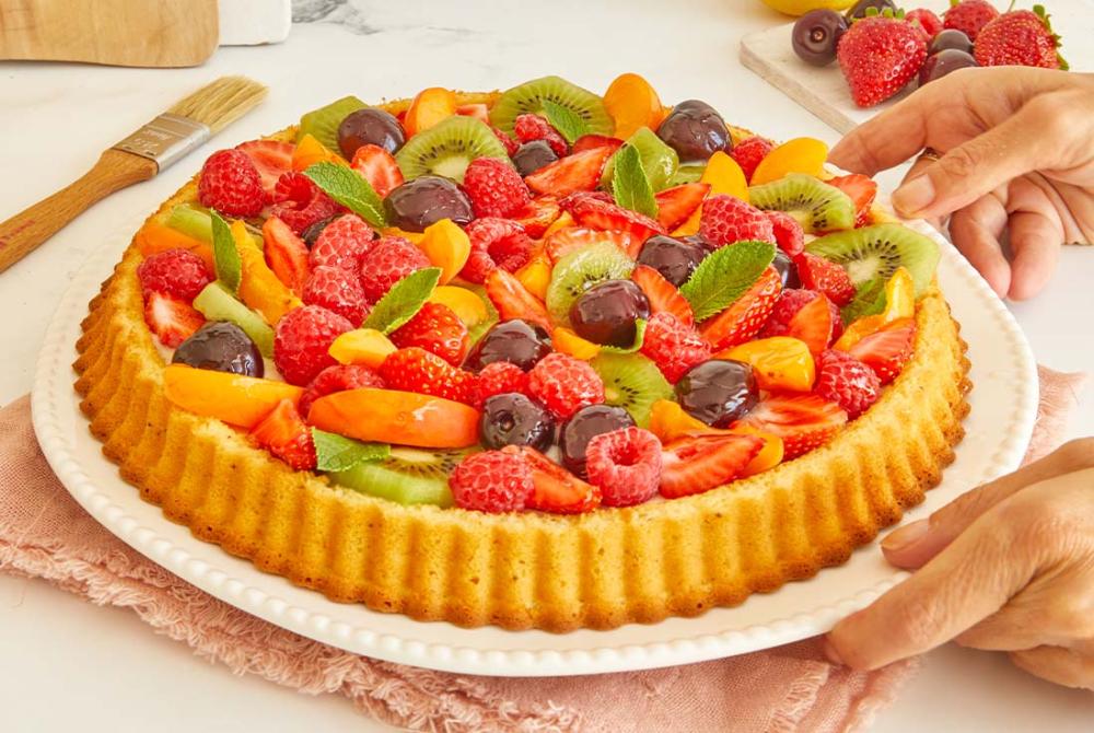 Tarte aux fruits frais décorée sur un plat blanc Tarte aux fruits frais maison composée de fraises, framboises, kiwis et cerises, nappée de brillant et décorée de menthe fraîche, posée sur une assiette à dessert.