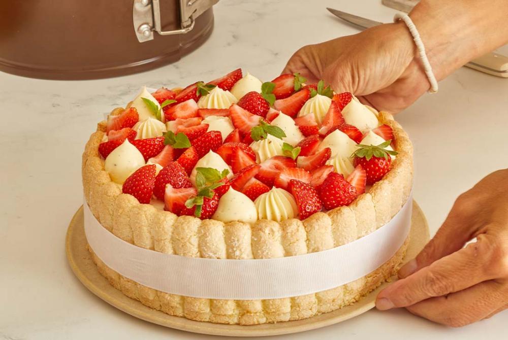 Une personne ajuste une charlotte aux fraises maison sur une assiette dans une cuisine, avec des biscuits &agrave; la cuill&egrave;re autour du g&acirc;teau et des fraises fra&icirc;ches en garniture.
