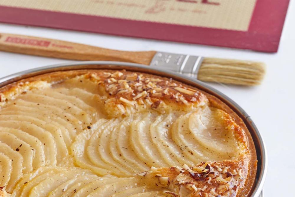 Image d'une tarte bourdaloue aux poires et aux amandes avec son cercle pâtissier, posée sur un plan de travail avec un pinceau de pâtisserie et un tapis de cuisson posés à côté