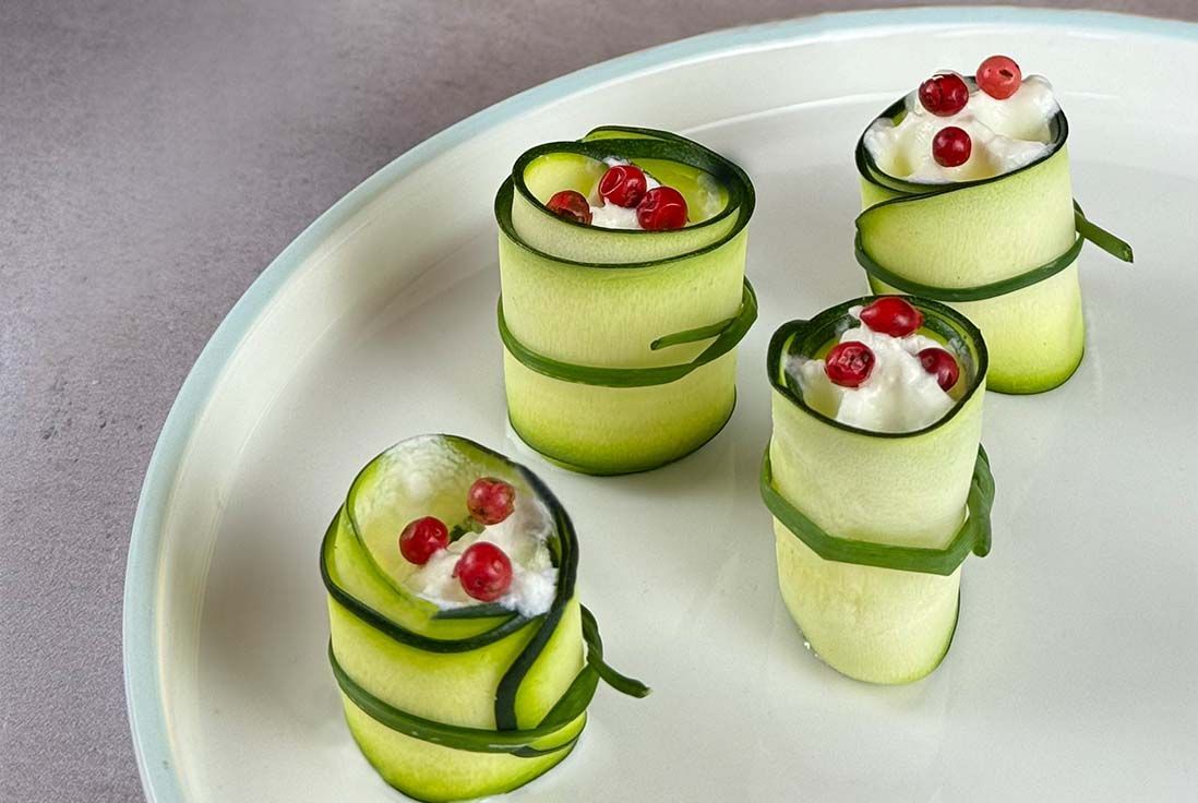 Quatre amuse-bouches en forme de rouleaux, composés de fines tranches de courgette garnies de crème blanche et surmontées de baies roses, sur une assiette