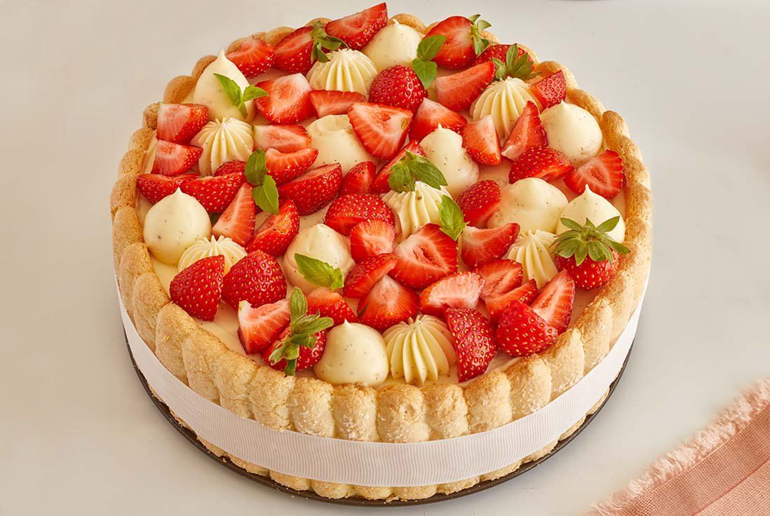 Une charlotte aux fraises maison posée sur une table, garnie de crème pâtissière pochée, de quartiers de fraises fraîches et de petites feuilles de menthe, entourée de biscuits à la cuillère maintenus par un ruban blanc
