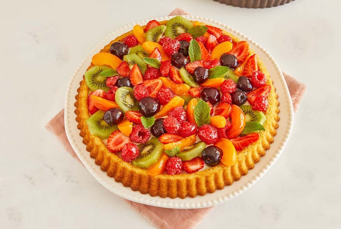 Tarte aux fruits colorée sur un fond blanc, garnie de fraises, framboises, kiwis, abricots et cerises, décorée de feuilles de menthe fraîche, avec un pinceau de cuisine à côté.