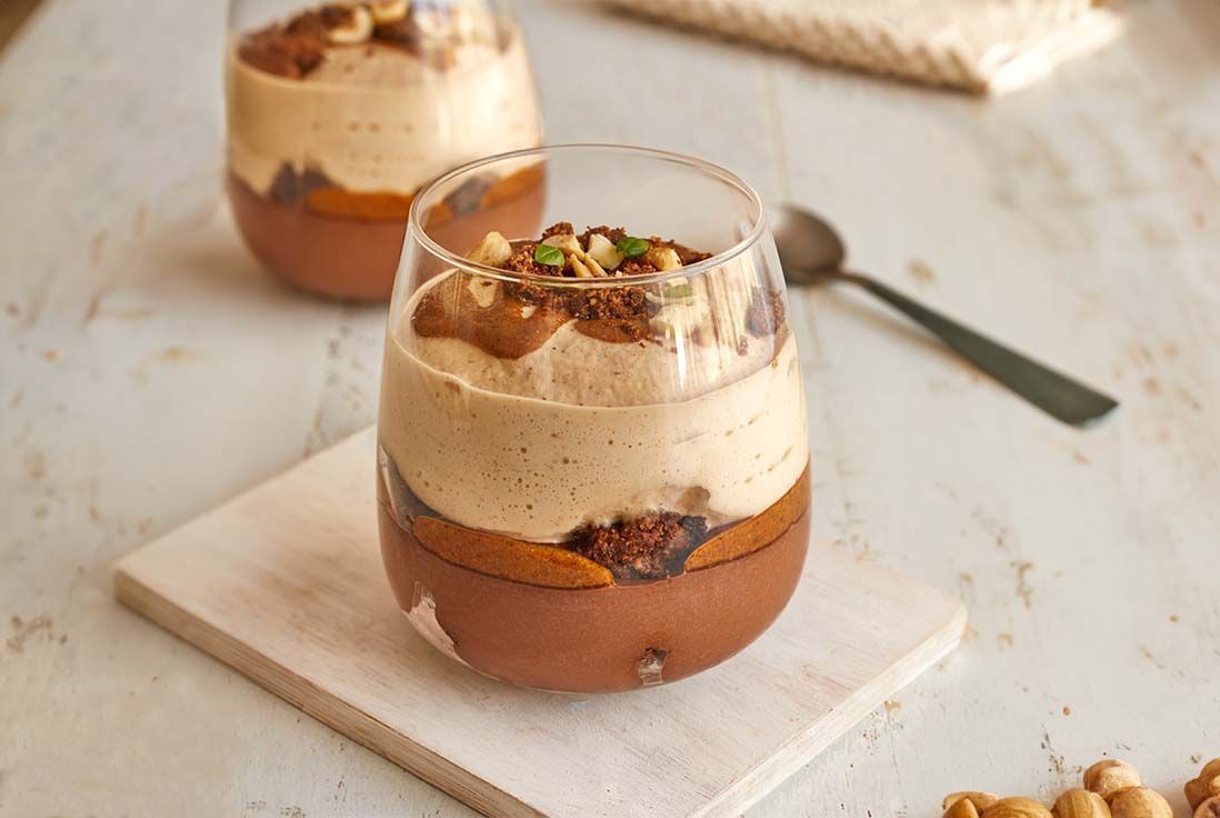 Deux verrines gourmandes au chocolat et praliné, posées sur une planche en bois. Une cuillère se trouve en arrière-plan sur une surface en bois clair.